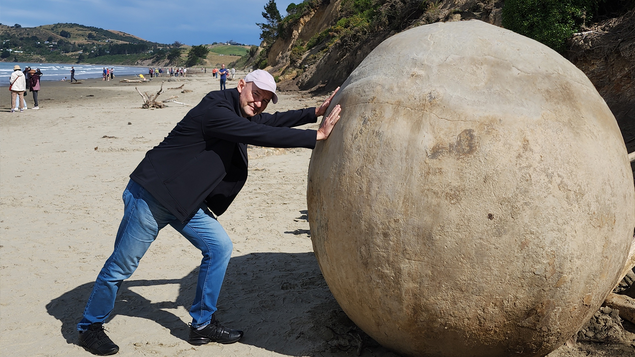 Турист позирует рядом с огромным валуном Моераки на пляже